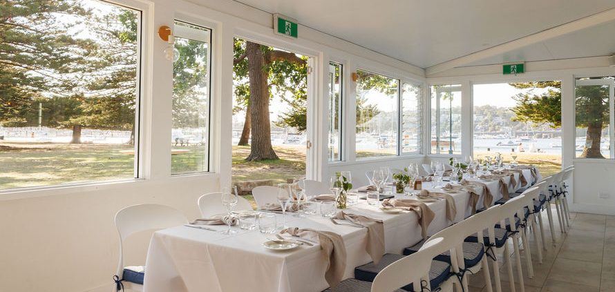 Bosk's glassed-in dining room at Clontarf, a long table set with white linen, the reserve's Moreton Bay figs and the moored boats of Middle Harbour visible through the windows.