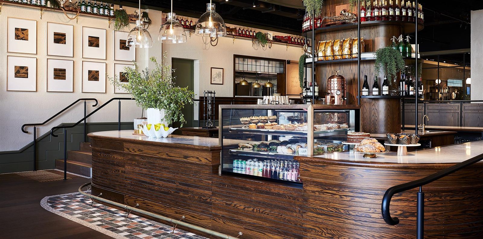L'americano's curved dark-timber counter with a glass pastry cabinet of croissants, sandwiches and cakes, brass fittings, glass pendant lamps and a mosaic-tiled floor.