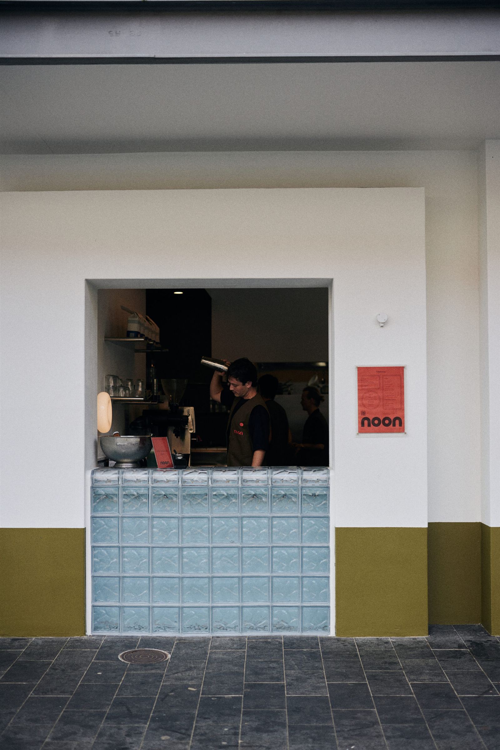 Noon's street-facing service window on Raglan Street: a barista working the espresso machine behind a pale pressed-tin counter, a red Noon poster on the white wall beside it.