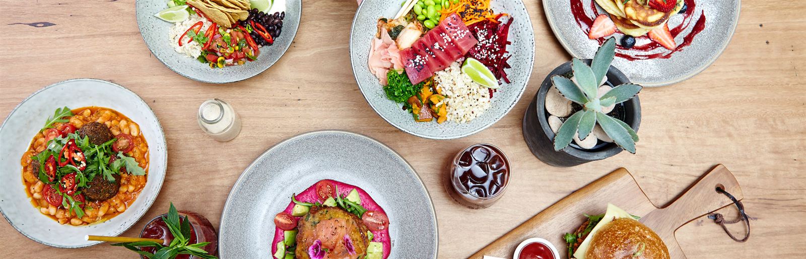 Brunch spread on a timber table at Roseberry Street Cafe: falafel with white beans and rocket, a tuna poke bowl, a corn fritter stack on beetroot puree, pancakes with berry coulis, an iced coffee and a chicken burger on a board.