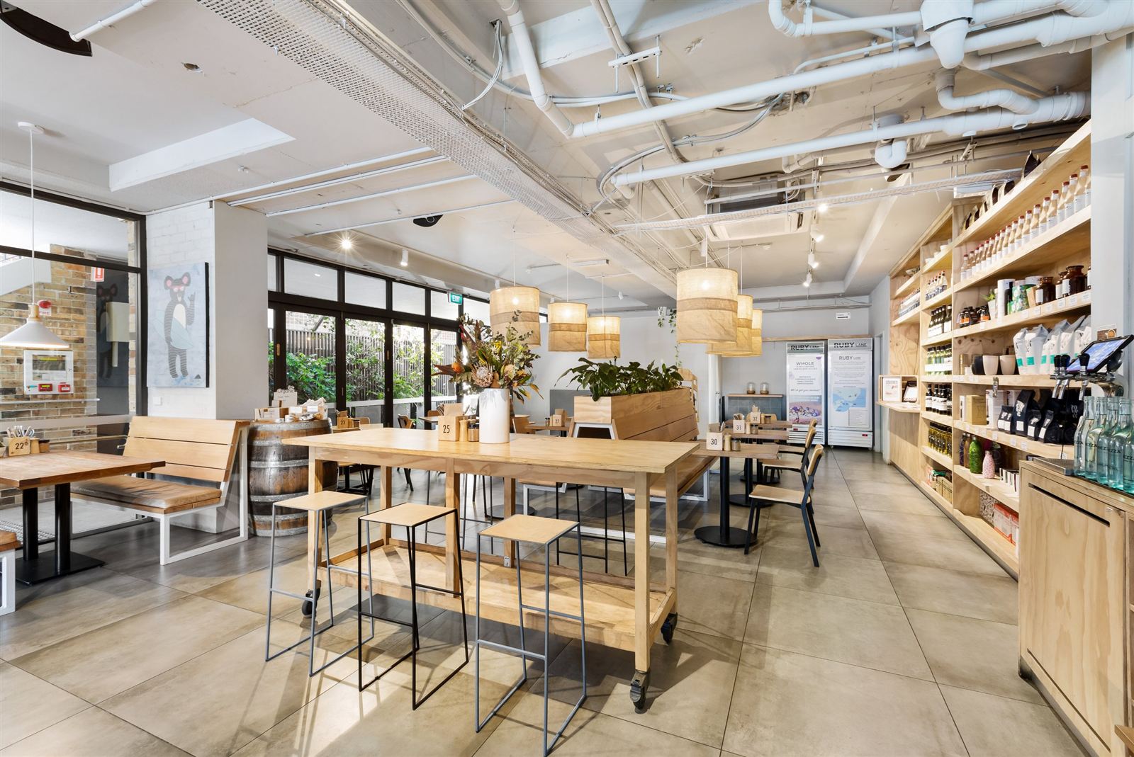 The interior of Ruby Lane Wholefoods: a long timber communal table with stools, woven pendant lights, polished concrete floor, and floor-to-ceiling shelves of oils, jars and pantry goods along the right-hand wall.