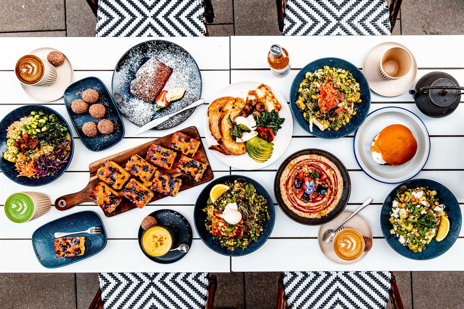 Flat-lay of a long white café table set with brunch: poached eggs on sourdough with halloumi and avocado, grain bowls with edamame and salmon, blueberry slice on a wooden board, a matcha latte and two flat whites with rosetta art.